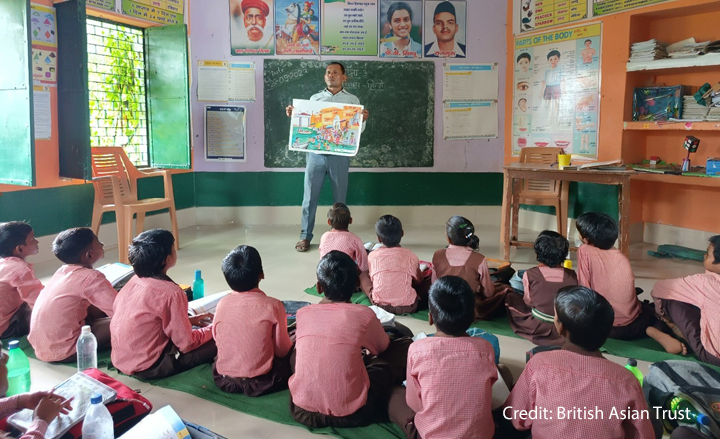 Measuring What matters A male teacher stands at the front of a class with students sitting on mats on the floor, India.