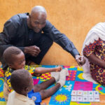 A male community member plays with young children sitting on a mat, overseen by a female community member.