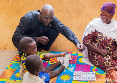 A male community member plays with young children sitting on a mat, overseen by a female community member.