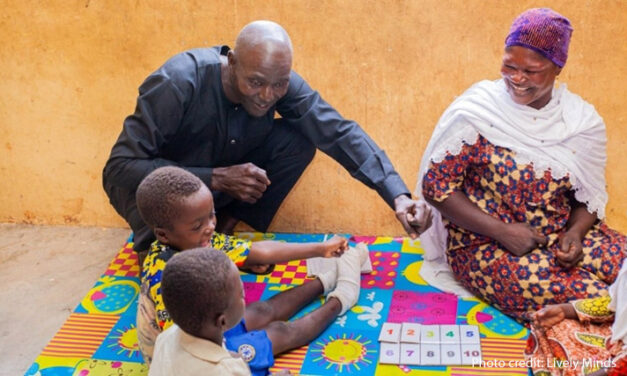 A male community member plays with young children sitting on a mat, overseen by a female community member.