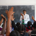 Male teacher stands at the front of a crowded classroom with hands raised, India.