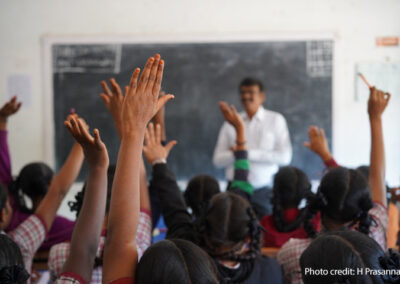 Male teacher stands at the front of a crowded classroom with hands raised, India.