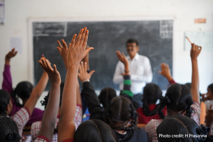 Male teacher stands at the front of a crowded classroom with hands raised, India.