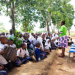 Female teacher stands in front of an outdoor class under trees while the students sit on the ground.