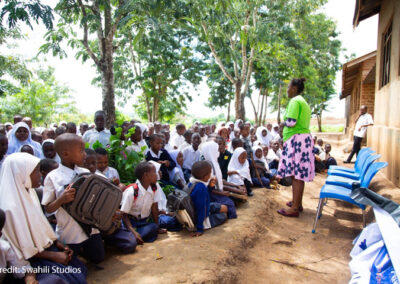 Female teacher stands in front of an outdoor class under trees while the students sit on the ground.