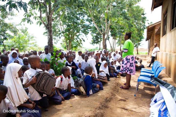 Educating under strain Female teacher stands in front of an outdoor class under trees while the students sit on the ground.