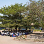 Primary school students wait for their class to start in the shade of a tree, Tanzania.