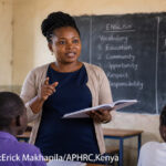 Female teacher at the front of a class with a book in her hand. Blackboard at the rear with English lesson points