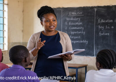 Female teacher at the front of a class with a book in her hand. Blackboard at the rear with English lesson points