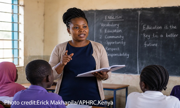 Refugee teachers have skills Female teacher at the front of a class with a book in her hand. Blackboard at the rear with English lesson points
